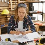 Hardworking young hispanic woman carpenter joyfully counting cash dollars in her indoor carpentry workshop, basking in the satisfaction of her successful woodwork business